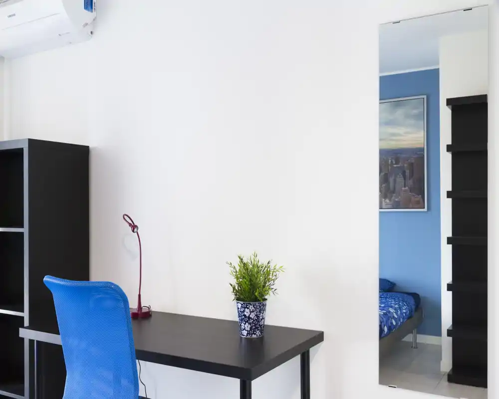 A bright, tidy bedroom corner shown via a workspace in the foreground; a bed is visible in the mirror reflection at right. The scene features a modern desk, blue chair, and decorative plant, with clean white walls and good natural light.