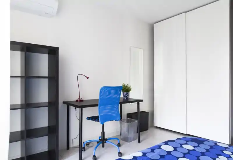 Bright, modern bedroom corner with a blue-patterned bed cover, a compact desk with a blue swivel chair, wardrobe and shelving—clean, minimal and well lit for a comfortable stay.