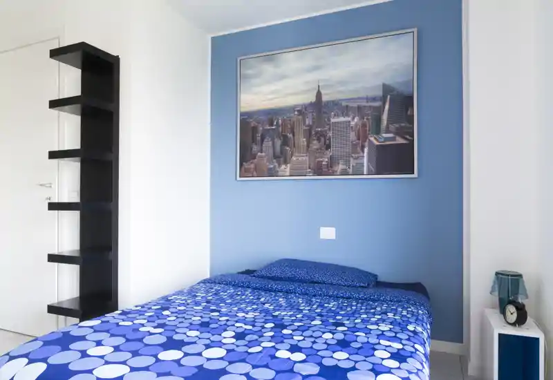 Bright, neatly staged bedroom with a double bed against a blue accent wall and a framed cityscape. The room feels fresh and well lit, with coordinated blue bedding and simple shelving.