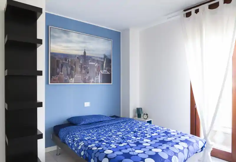 Bright, tidy bedroom with a blue accent wall and a double bed dressed in patterned blue bedding. Natural light fills the room through sheer curtains, and a framed cityscape and bedside accessories give a polished, welcoming feel.