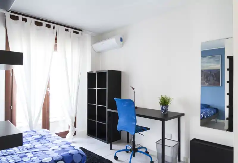 Bright, modern bedroom with a patterned blue bedspread, desk and shelving—well lit by a large window and neatly staged for rental photos.