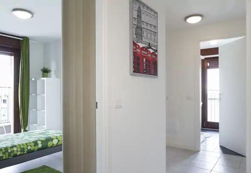 Bright modern bedroom with a patterned green bedspread, tall window/door to a balcony, and minimal white shelving — tidy and well lit for rental photos.