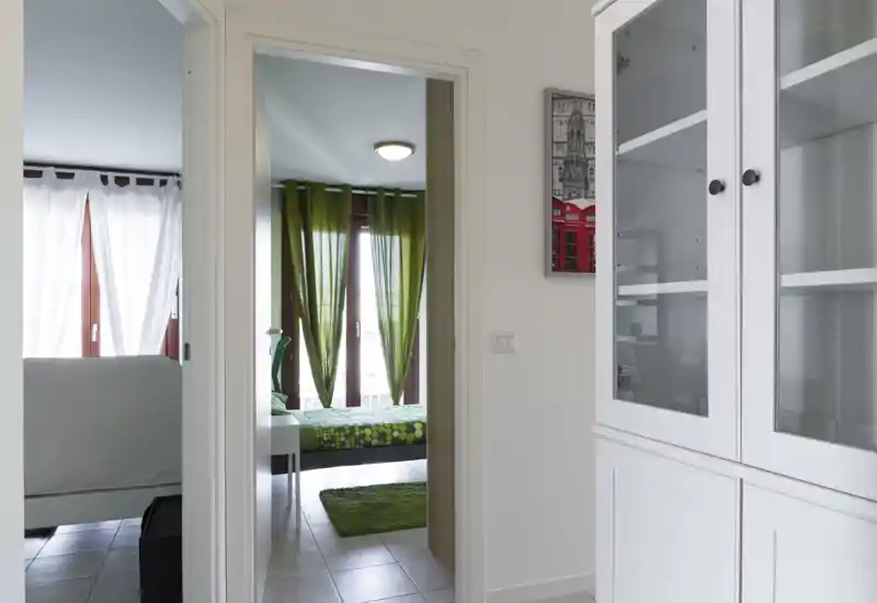 Bright, modern bedroom visible through a doorway with green curtains, patterned bedding and a small matching rug—clean lines and plenty of natural light.