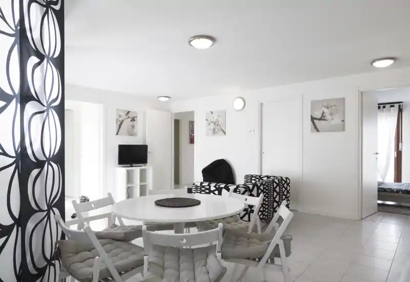 Bright, modern living area with a round dining table, cushioned folding chairs and patterned lounge seating; neutral white walls and contemporary black-and-white decor create a clean, airy space.