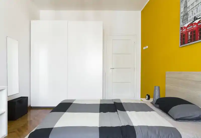 Dormitorio moderno y luminoso con cama grande en primer plano, armario blanco y pared de acento amarilla. Líneas limpias y luz natural crean un espacio fresco y acogedor, ideal para anuncios de alquiler.