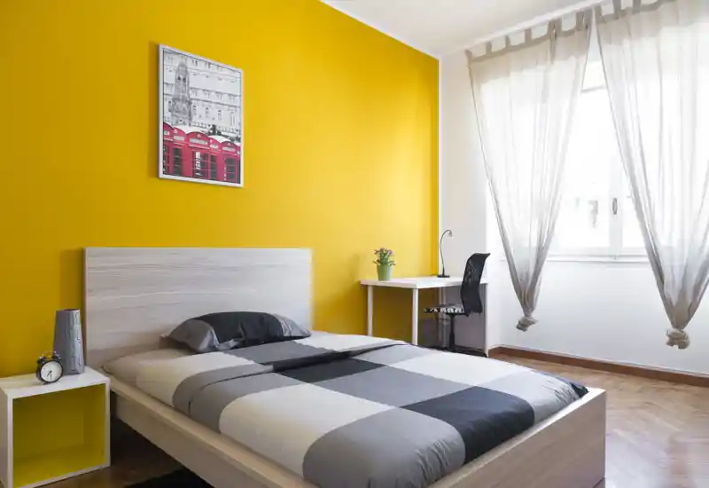 Dormitorio moderno y luminoso con cama de madera clara, edredón gris a cuadros y una pared amarilla de acento. Un pequeño escritorio junto a la ventana y cortinas translucidas completan el ambiente aireado.