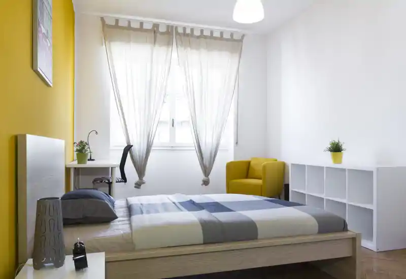 Dormitorio moderno y luminoso con cama doble, gran ventana que aporta luz natural, pared amarilla de acento y sillón acogedor. Líneas limpias y almacenamiento sencillo, perfecto para alquileres temporales.