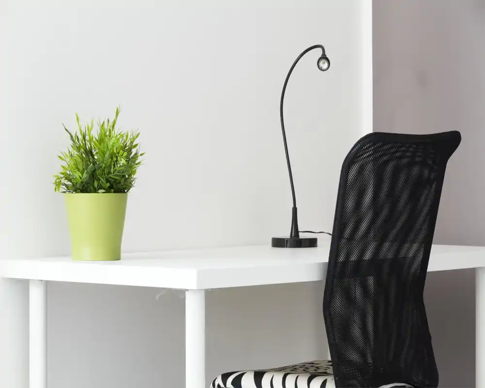 Angolo scrivania minimal con tavolo bianco, sedia in rete nera e una piccola lampada; una pianta in vaso verde aggiunge un tocco fresco in uno spazio di lavoro luminoso e ordinato.