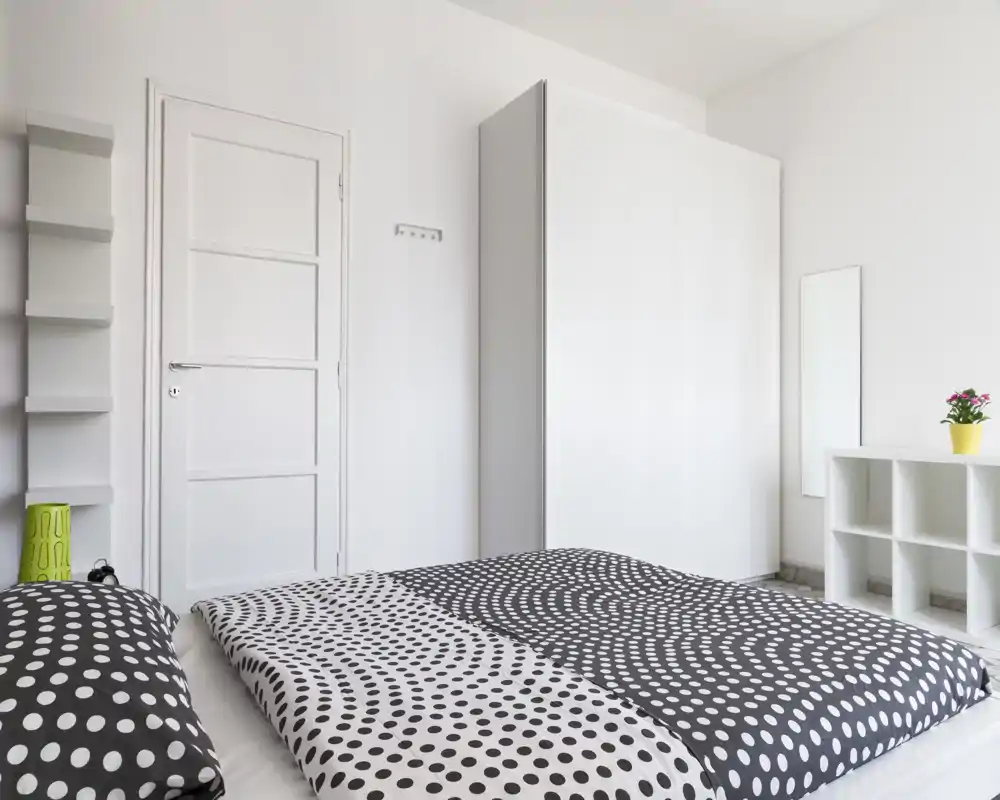 Camera da letto luminosa e moderna con un grande letto basso con copriletto a pois, armadio bianco e scaffalature minimal — spazio pulito e arioso ideale per annunci.