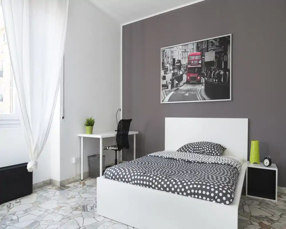 Camera da letto luminosa e moderna con letto bianco, biancheria a pois e una scrivania vicino alla finestra. Luce naturale e arredi essenziali creano un'atmosfera fresca e accogliente, perfetta per affitti.