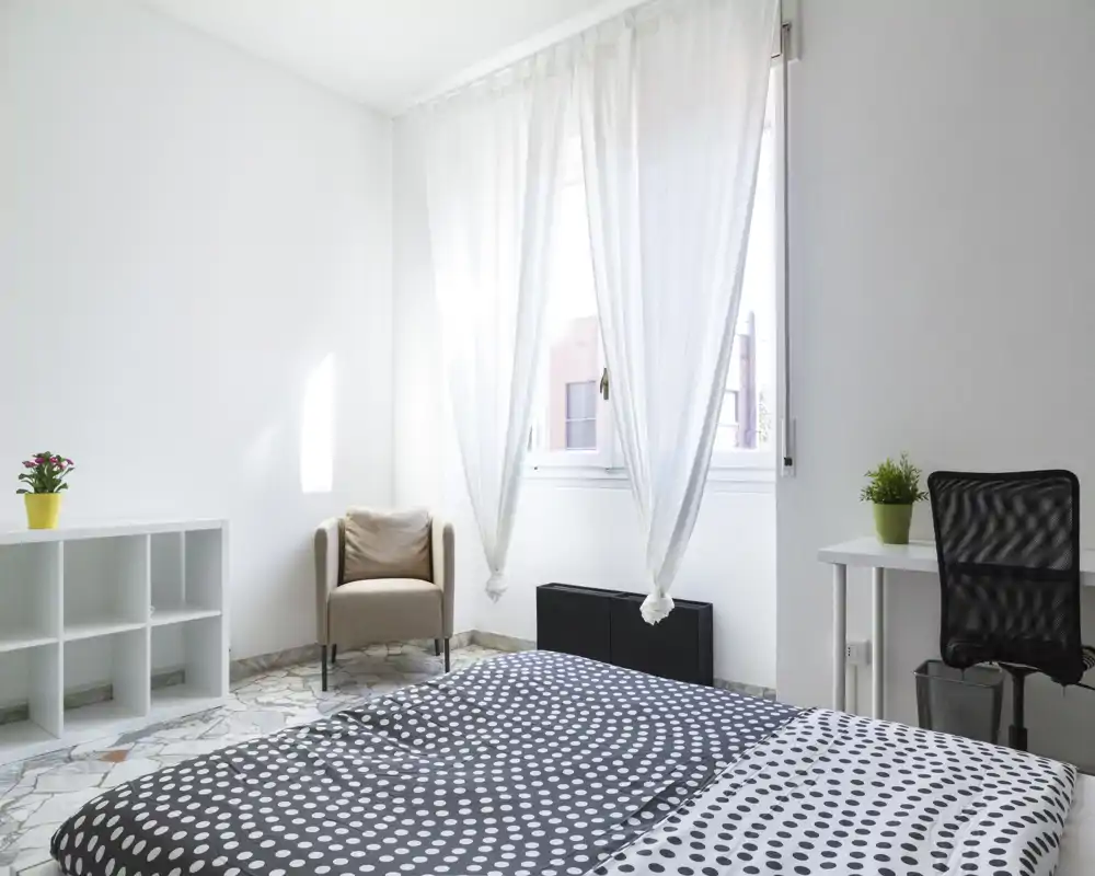 Camera da letto luminosa e minimalista con un grande letto in primo piano, tende leggere che lasciano entrare molta luce naturale, una poltrona comoda e mensole semplici per riporre.
