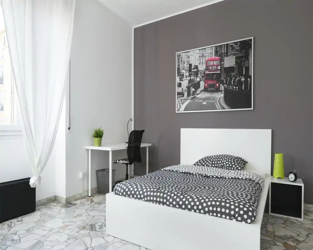 Camera da letto moderna e luminosa con letto bianco, copriletto a pois, una scrivania e sedia vicino alla finestra e una stampa urbana sopra la testiera.