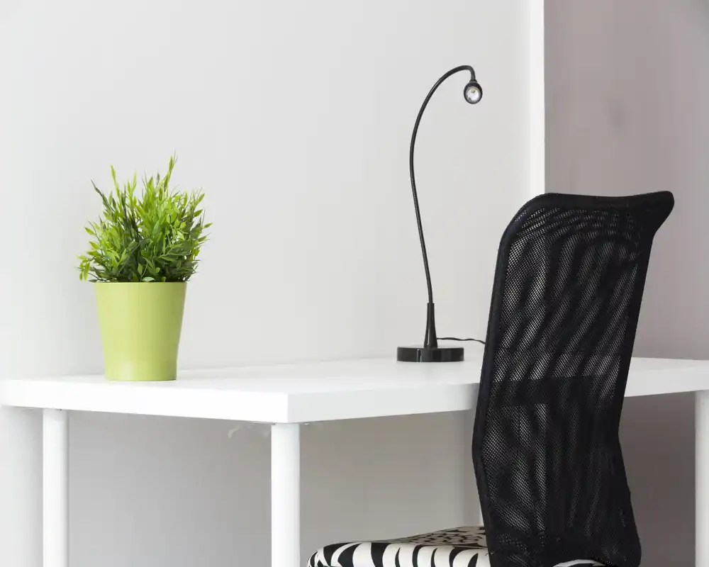Angolo lavoro luminoso e minimale con scrivania bianca, sedia in rete nera, lampada da tavolo e una pianta in vaso — perfetto per studiare o lavorare da casa.