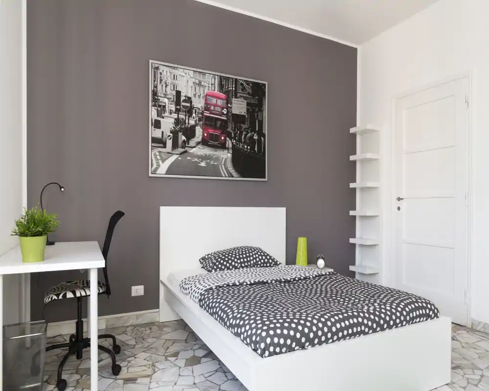 Camera da letto moderna e luminosa con letto bianco, biancheria a pois, una scrivania e una sedia, e un quadro decorativo. Ambiente ordinato e piacevole, adatto per annunci di affitto.