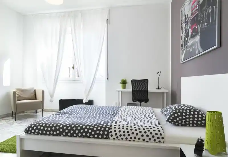 Camera da letto moderna e luminosa con letto matrimoniale coperto da biancheria a pois, una scrivania con sedia vicino alla finestra e una poltrona nell'angolo. Pareti bianche e luce naturale conferiscono un'atmosfera ariosa.