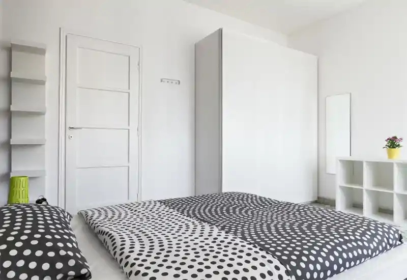 Camera da letto minimal e luminosa con un letto basso matrimoniale con copripiumino a pois bianco e nero, armadio bianco, mensole e una piccola pianta. L’ambiente è pulito, ben illuminato e moderno — adatto per annunci.
