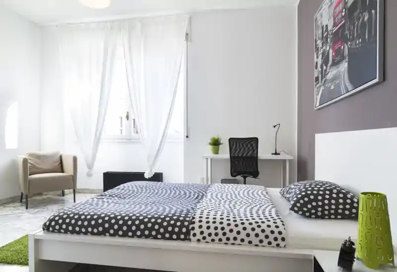 Camera da letto luminosa e moderna con letto bianco ampio con biancheria a pois, scrivania e sedia vicino alla finestra e una poltrona nell'angolo. Luce naturale abbondante e arredo minimal rendono l'ambiente accogliente e ideale per annunci.