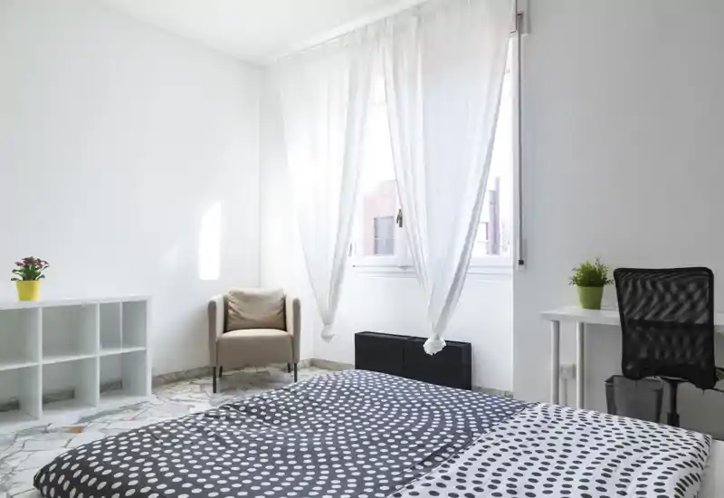 Dormitorio luminoso y minimalista con una gran cama en primer plano, cortinas translúcidas que dejan pasar abundante luz natural, un sillón cómodo y estanterías sencillas.