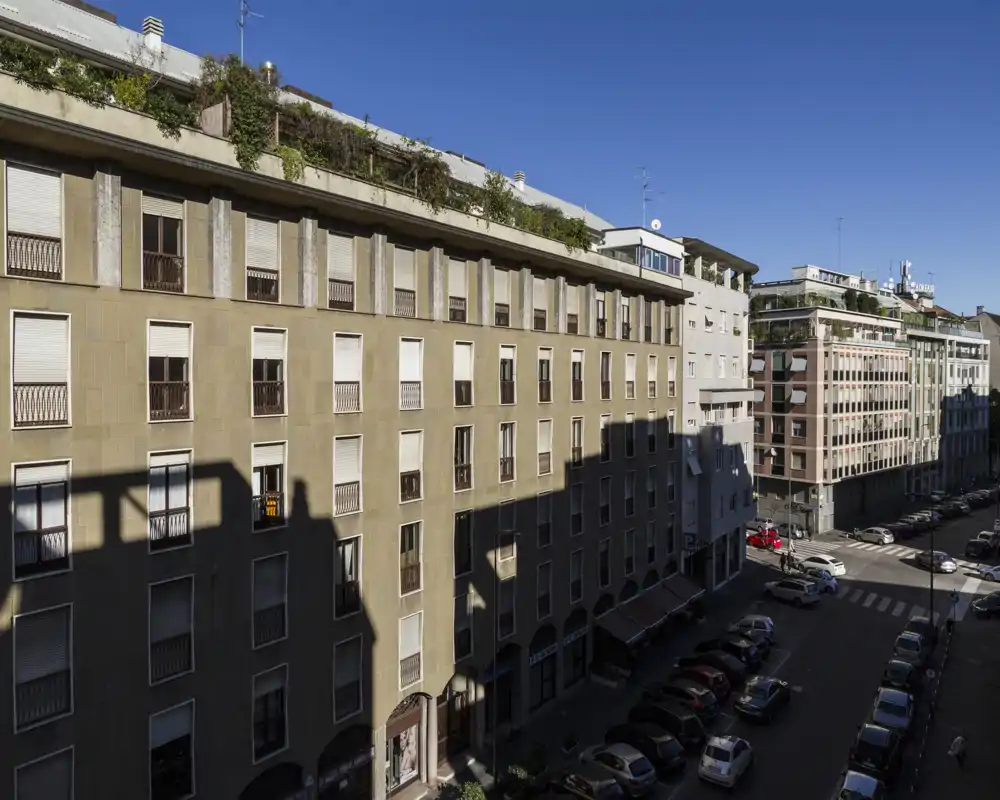 Veduta di strada e palazzi da un balcone o finestra: scena urbana soleggiata con auto parcheggiate, edifici residenziali e cielo sereno.
