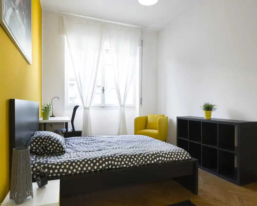 Camera da letto moderna e luminosa con piumone a pois, struttura del letto bassa nera, parete d'accento gialla e poltrona coordinata; finestra ampia con tende leggere che diffondono luce naturale.