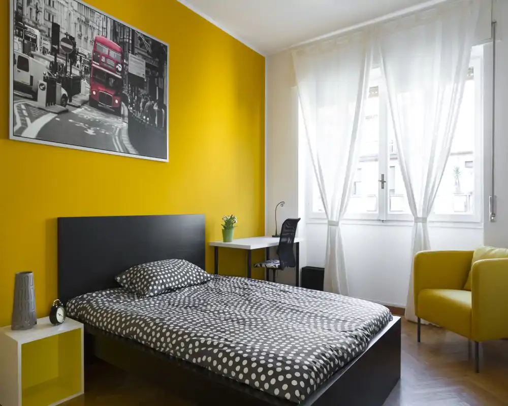 Camera da letto moderna e luminosa con letto a pois, parete accentuata gialla e ampia finestra che garantisce molta luce naturale. Una scrivania e una poltrona completano l’arredamento funzionale.