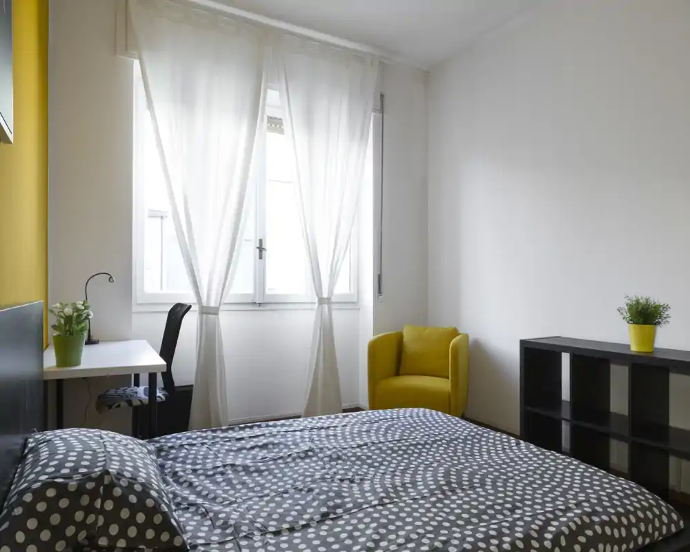 Camera da letto moderna e luminosa con letto matrimoniale con copripiumino a pois, una scrivania vicino alla finestra e una poltrona gialla come tocco di colore.