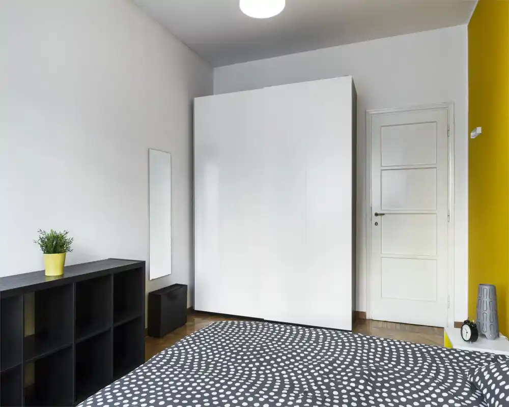 Camera da letto minimalista con copriletto a motivo, grande armadio bianco e scaffale nero; luminosa, ordinata e moderna con parete d'accento gialla.