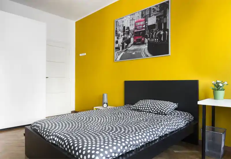 Camera da letto moderna e luminosa con letto singolo dalla struttura nera, biancheria a pois e una parete gialla a contrasto con un quadro urbano.