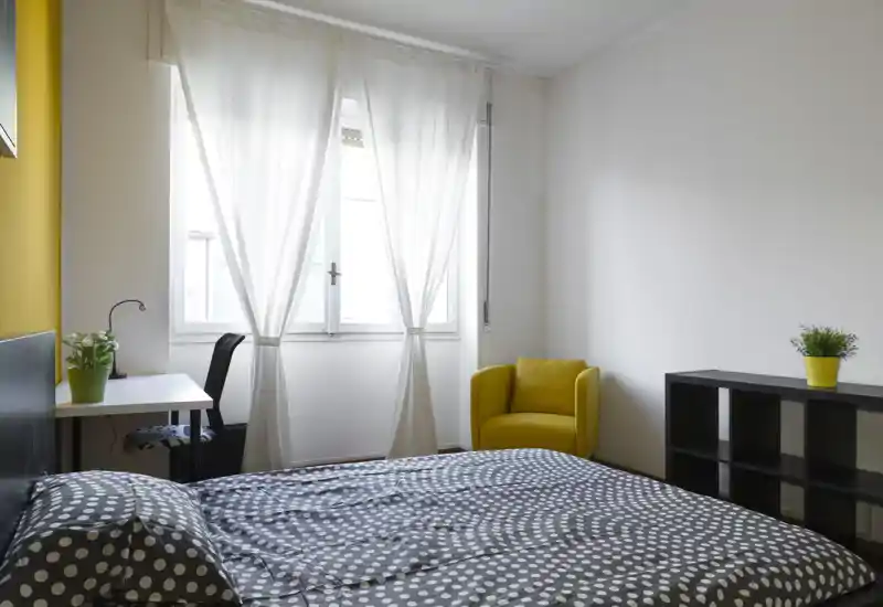 Camera da letto moderna e luminosa con letto matrimoniale con copripiumino a pois, una scrivania vicino alla finestra e una poltrona gialla come tocco di colore.