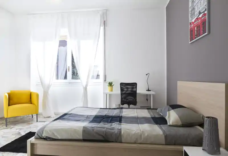 Dormitorio moderno y luminoso con cama doble, cabecero de madera clara, escritorio junto a la ventana y un sillón amarillo — estilo minimalista y mucha luz natural.