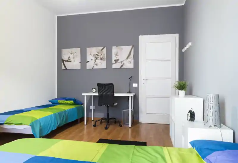 Bright, modern bedroom with two single beds dressed in colorful blue and green linens, a white desk with ergonomic chair, wall art and wooden flooring — ideal for shared or student accommodation.
