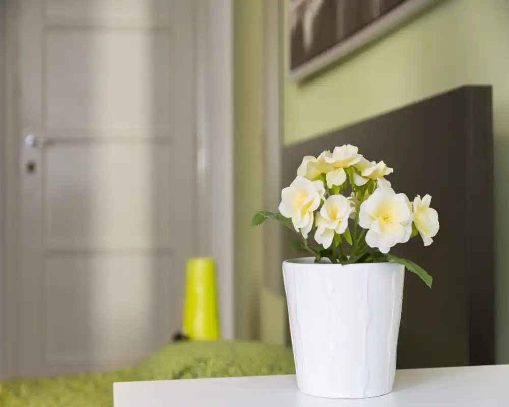 Gros plan d’un coin de chambre avec un pot de fleurs blanc sur la table de chevet, une tête de lit sombre et des accents verts pour une atmosphère moderne et apaisante.