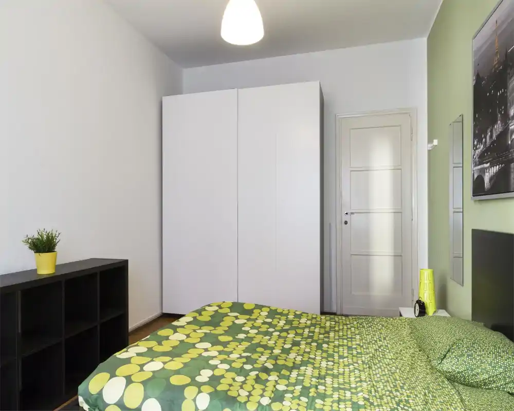 Chambre moderne et lumineuse avec dessus-de-lit vert à motifs, armoire blanche et mobilier minimal. La pièce est bien éclairée et décorée d'une petite plante et d'accents assortis pour une atmosphère fraîche et accueillante.