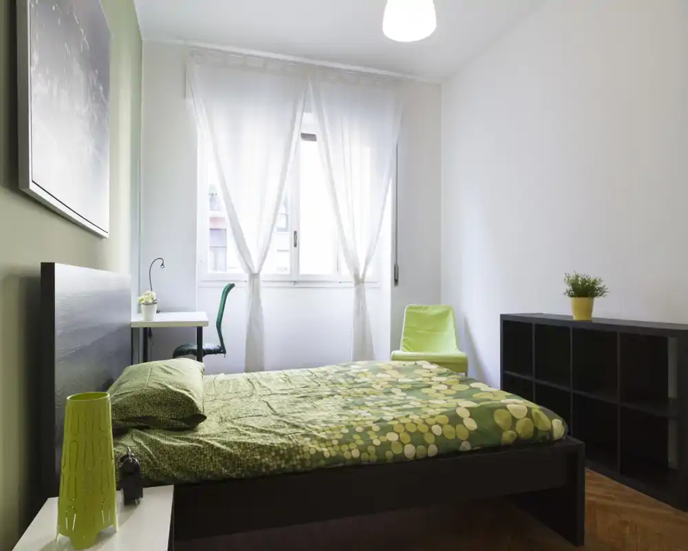 Chambre moderne et lumineuse avec un lit double habillé d'une couette verte à motifs, un petit bureau près de la fenêtre, une chaise et une étagère modulable. Grande fenêtre avec voilages apportant une lumière naturelle abondante.