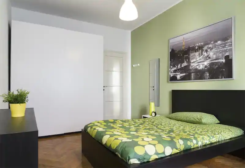Chambre moderne et lumineuse avec un lit double habillé d’une couette verte à motifs, une grande armoire blanche et une décoration minimaliste. Le parquet chaleureux et une photo de ville au-dessus de la tête de lit créent une ambiance fraîche et accueillante.