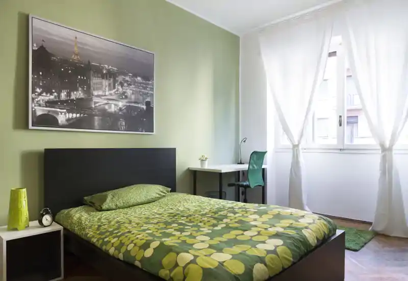 Chambre moderne et lumineuse avec un lit double habillé d'une couette verte à motifs, une tête de lit noire, une table de chevet et un bureau près d'une grande fenêtre laissant entrer beaucoup de lumière naturelle.