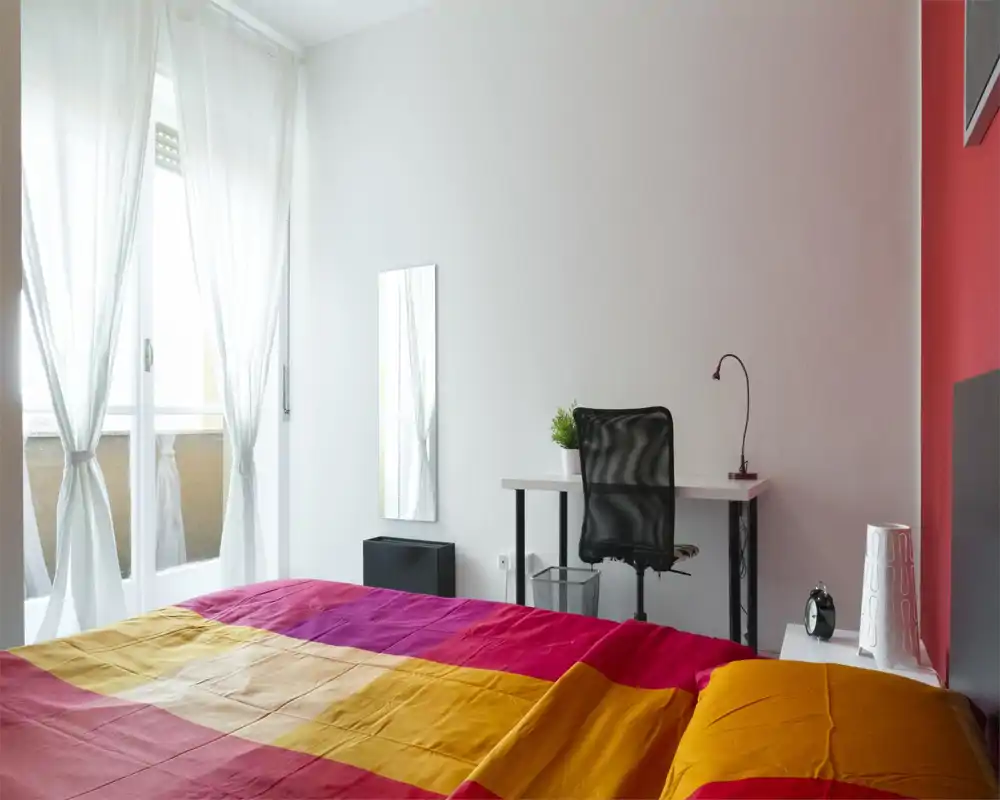 Sunny, modern bedroom with colorful bedding, a desk and chair by the wall, and sheer curtains letting in soft natural light.