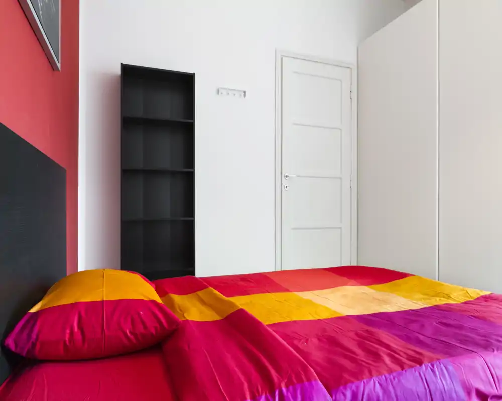 Bright, colorful bedroom with a large bed covered in red, orange and pink linens; simple shelving and a white door complete the minimalist layout.