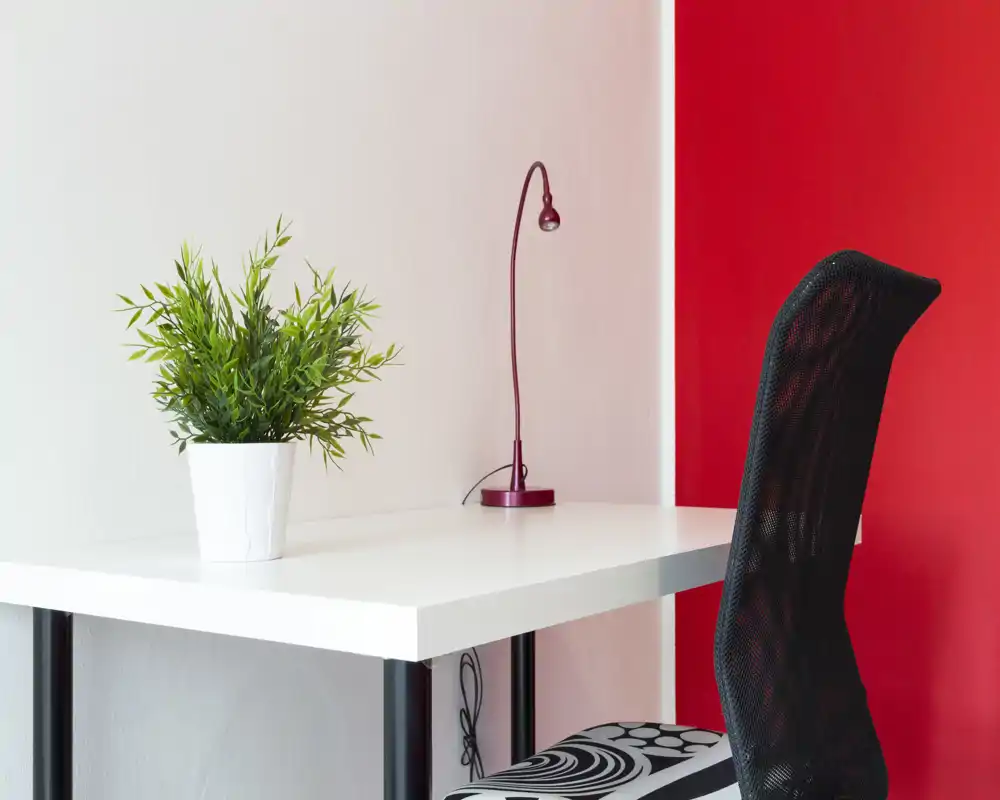 Bright, modern workspace corner with a white desk, black mesh chair, a red desk lamp and a small potted plant — minimal and stylish.