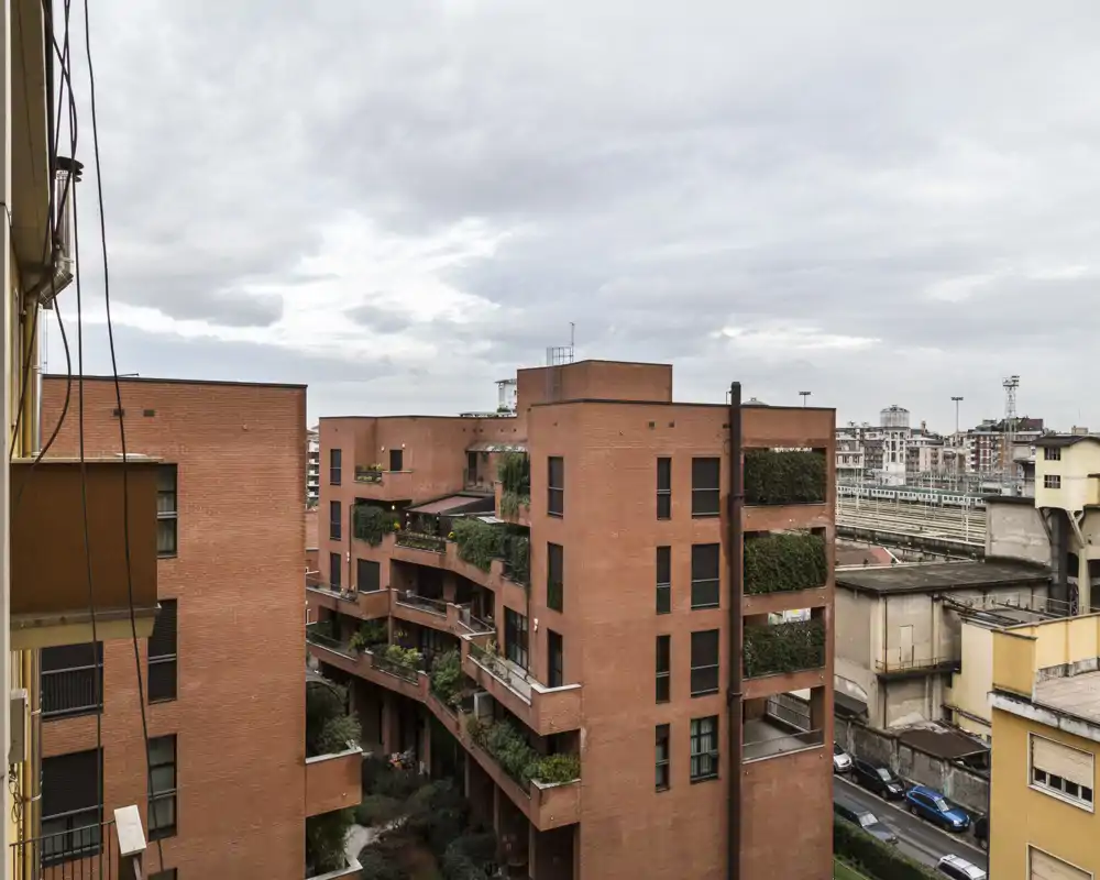 Exterior view of a red-brick residential building with planted balconies and an urban streetscape under a cloudy sky — suitable for showing the building facade and neighborhood context.