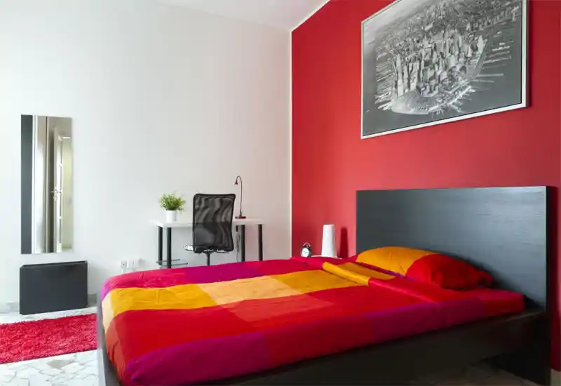 Bright, modern bedroom with a large double bed dressed in colorful red and orange linens, a black headboard, and a small desk with chair — stylish and ready for guests.