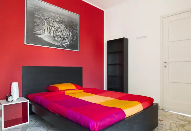 Bright, colorful bedroom with a low black bedframe and vibrant multi-color bedding against a striking red accent wall; includes a bedside table, alarm clock and tall shelving unit.