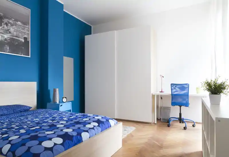 Bright, modern bedroom with a patterned blue bedspread, large white wardrobe and a small desk by the window. The room is well lit with natural light and has a clean, contemporary styling ideal for a rental listing.