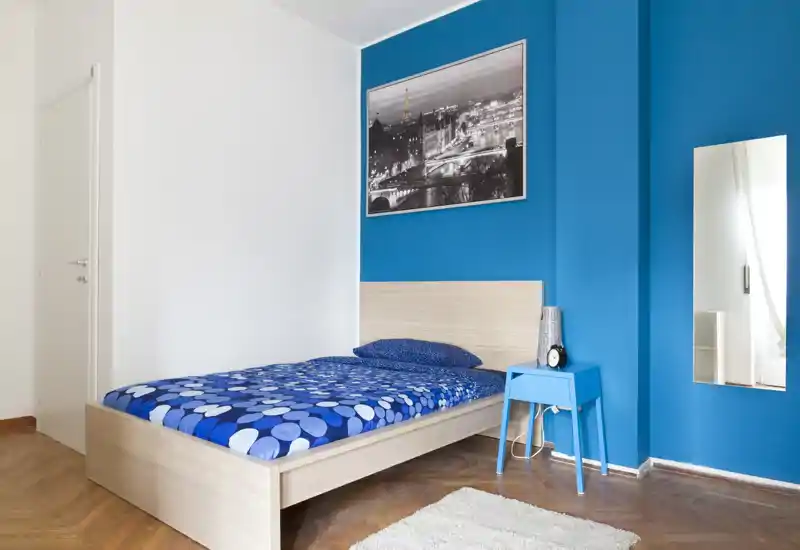 Bright, modern single bedroom with a light wood bed frame, patterned blue bedding, matching bedside table and a decorative rug — clean, contemporary styling with strong blue accent wall.