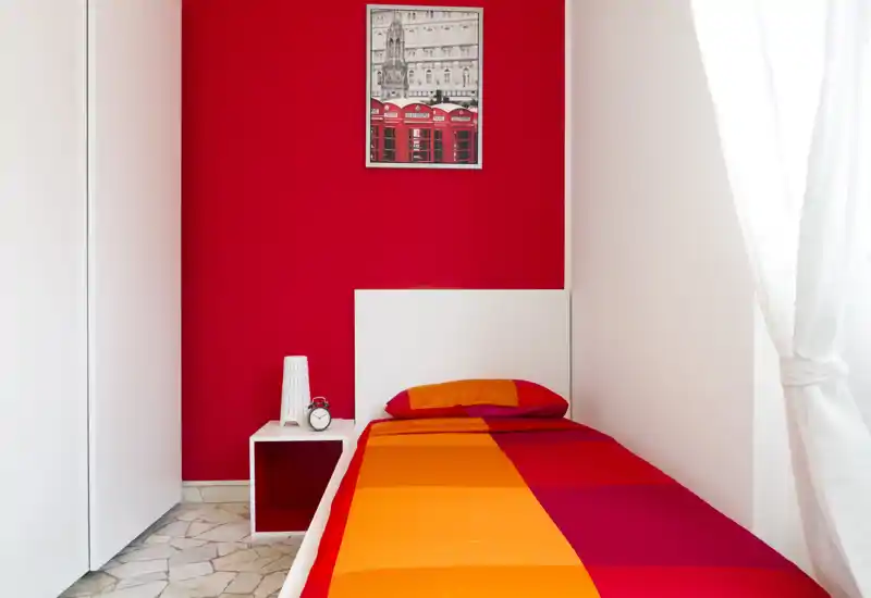 Bright single bedroom with a colorful orange-and-red bed and a simple white headboard; natural light filters through sheer curtains and a small bedside shelf holds a lamp and alarm clock.