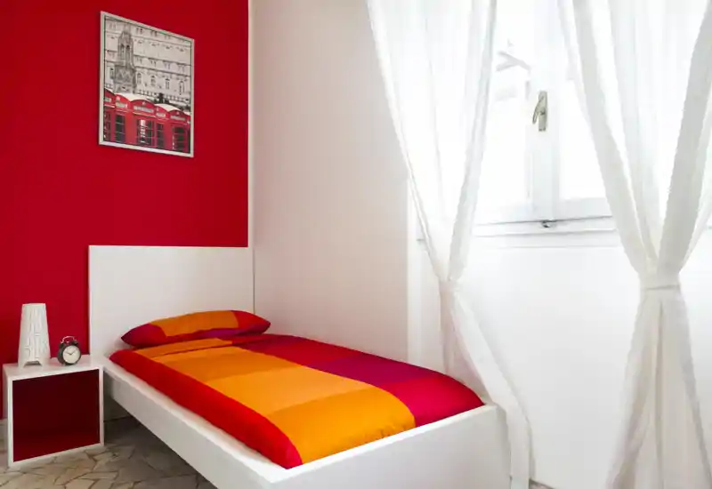 Bright single bedroom with a colorful orange-and-red duvet, simple white bed frame, bedside table and large window with sheer curtains letting in lots of natural light.