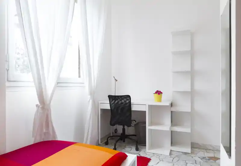 Bright, minimalist bedroom with a colorful bedspread, a compact white desk and shelving, and a window with sheer curtains providing abundant natural light.
