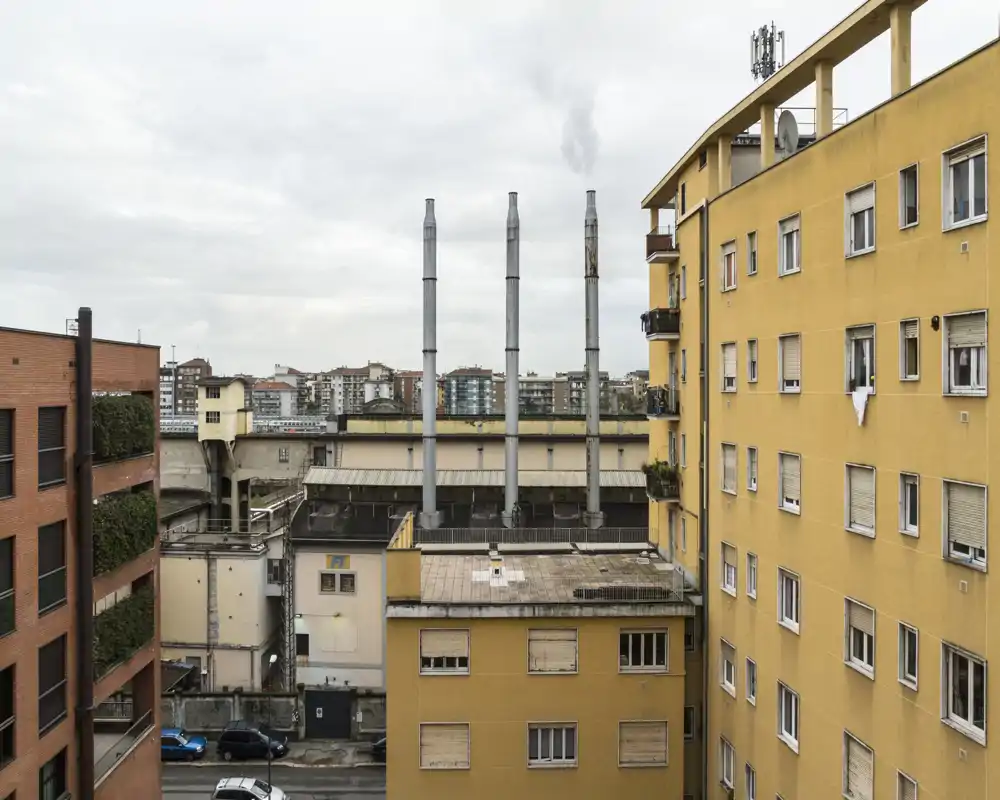 Vista esterna urbana con palazzi adiacenti, tetti e canne fumarie sotto un cielo coperto. Utile per mostrare l'ambiente e le linee di vista dell'edificio.