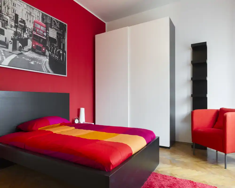 Camera da letto luminosa e moderna con letto basso in legno scuro, biancheria a righe colorate e una parete rossa di forte impatto. Armadio bianco e poltrona rossa completano l’ambiente accogliente.