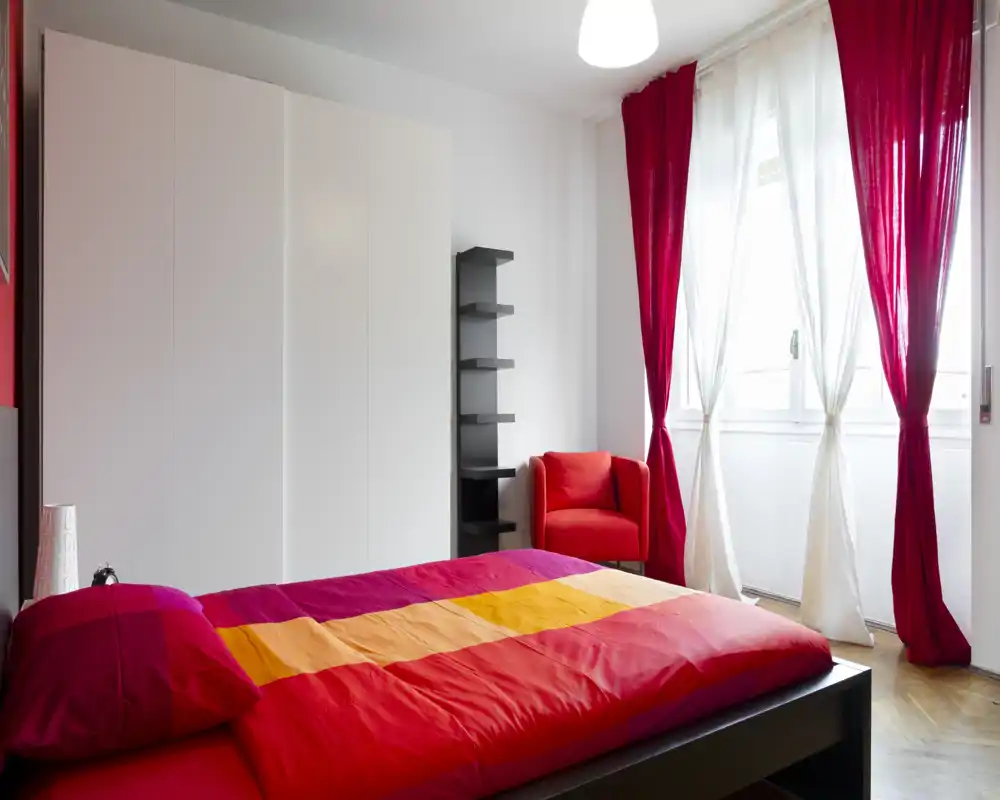 Camera da letto luminosa e colorata con un grande letto matrimoniale, poltrona rossa e finestra con tende rosse e bianche. Linee moderne e luce naturale abbondante, tessuti vivaci che rendono l’ambiente accogliente.